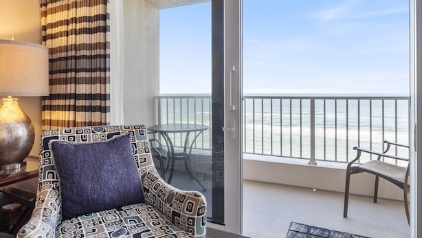 A balcony with a view of the beach, a patterned sofa, a small table, and a lamp. - Southpoint 707 La Casa Azul (Ponce Inlet)