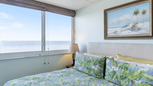 A bedroom with a bed, a window with a view of the ocean, a painting of a beach scene, and a nightstand with a lamp. - Southpoint 707 La Casa Azul (Ponce Inlet)