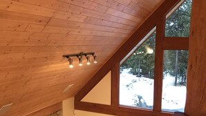 A cozy living room with a stone fireplace, wooden ceiling, and large windows offering a view of a snowy landscape. - Unbeatable Chalet at Base of Red Mountain (Rossland)