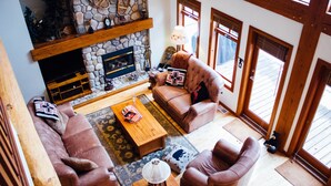 A cozy living room with a stone fireplace, leather sofas, a wooden coffee table, and a large window with wooden frames. - Unbeatable Chalet at Base of Red Mountain (Rossland)