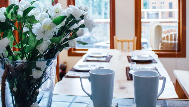 A dining table with two white ceramic cups, a vase with white flowers, and a wooden ceiling with a deer antler chandelier. - Unbeatable Chalet at Base of Red Mountain (Rossland)