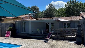 A wooden deck with a swimming pool, a covered patio area, and a small building with a tiled roof. - Near SOULAC/mer, air-conditioned house with heated indoor pool sleeps 4 (Grayan-et-l'Hôpital)