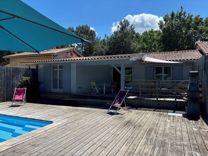 A wooden deck with a swimming pool, a covered patio area, and a small building with a tiled roof.