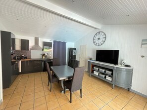 Uma cozinha compacta com eletrodomésticos modernos, uma sala de jantar com mesa e cadeiras, e uma televisão sobre um móvel.