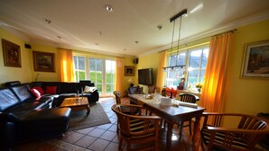 Sala de estar com sofá de couro preto, mesa de jantar de madeira e cadeiras. Há uma TV montada na parede e uma janela com cortinas.