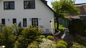 Uma casa branca de dois andares com telhado de cerâmica, numerada 29D, cercada por vegetação exuberante e um banco de pedra.