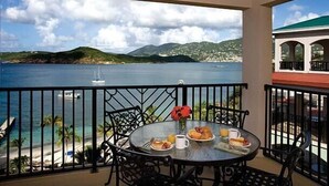 A balcony with a table set for breakfast overlooking a coastal view with mountains and a clear sky.