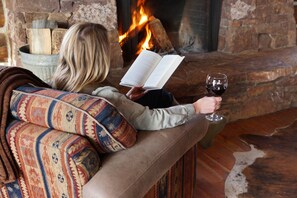 A person reading a book by a fireplace with logs and a glass of wine. - Cozy Two Bedroom Cabin on Private Montana Dude Ranch - Angler's Perch (Belt)