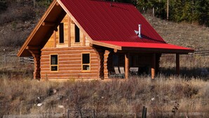 A log cabin with a red roof, a porch with chairs, and a window overlooking a grassy area. - Log Chalet with Mountain views and private lake (Lumby)