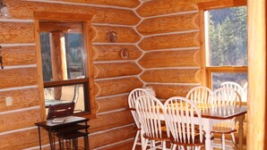 A log cabin interior with a wooden dining table and chairs, a window with a view of trees, and a wooden floor. - Log Chalet with Mountain views and private lake (Lumby)