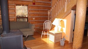 A cozy log cabin interior with a sofa, a white chair, a lamp, and a fireplace. - Log Chalet with Mountain views and private lake (Lumby)