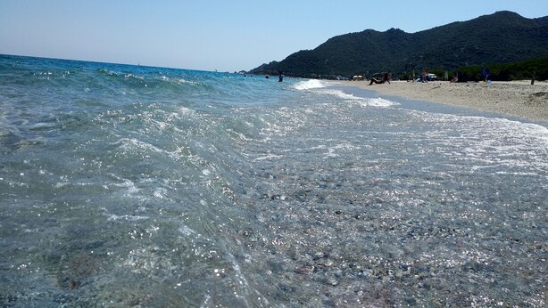 Ein Strand mit klarem Wasser, Kieselsteinen und einem Berg im Hintergrund.