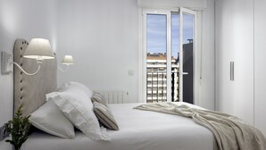 A bedroom with a bed, white walls, a radiator, a window with a view of buildings, and a door. - Always Easy I Ático Plaza (San Sebastián/Donostia)
