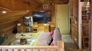 A cozy cabin interior with a wooden ceiling, a television mounted on the wall, a wooden bench, a cushioned sofa, and a small table with various items.