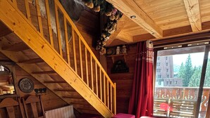 A cozy cabin interior with a wooden staircase, a sofa with patterned cushions, a coffee table with a box, and a view of a building through a window.