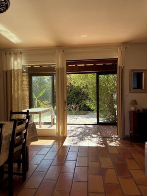 Habitación con suelo de baldosas, una mesa con sillas, un banco exterior y vistas a la vegetación a través de un ventanal.