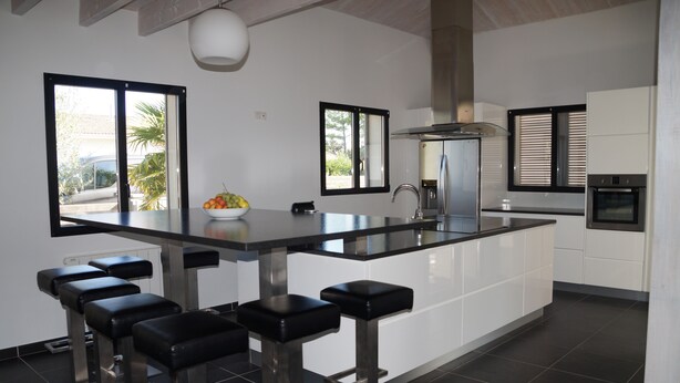 Una cocina moderna con una isla central, taburetes negros tipo bar, un cuenco de fruta y ventanas amplias.