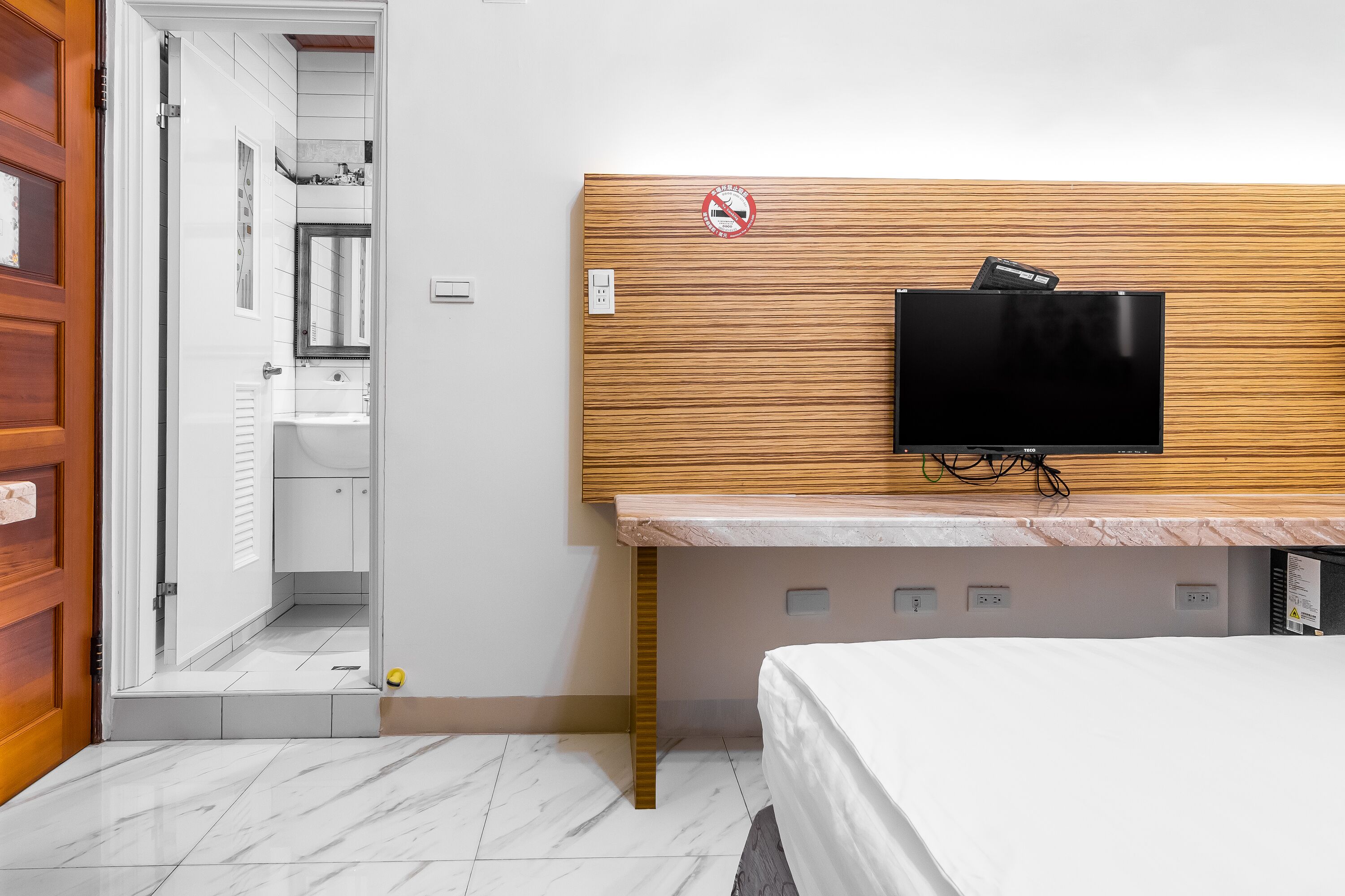 Une chambre d’hôtel avec une tête de lit en bois, un téléviseur à écran plat fixé au-dessus d’un comptoir en marbre, un lit avec une literie blanche et une salle de bain visible à travers une porte ouverte.