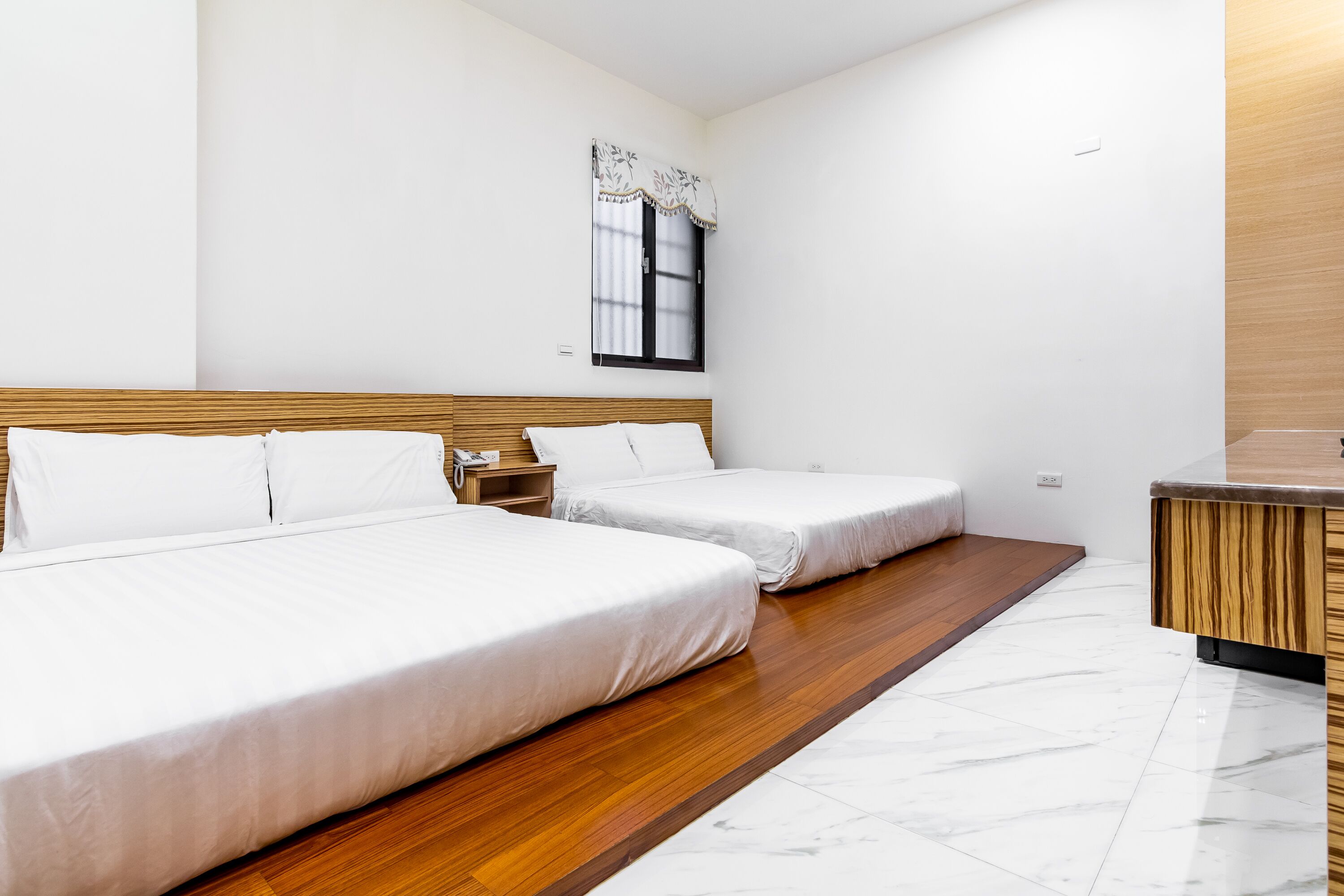 A hotel room with two beds, a wooden headboard, a nightstand, a window with a curtain, and a wooden counter.