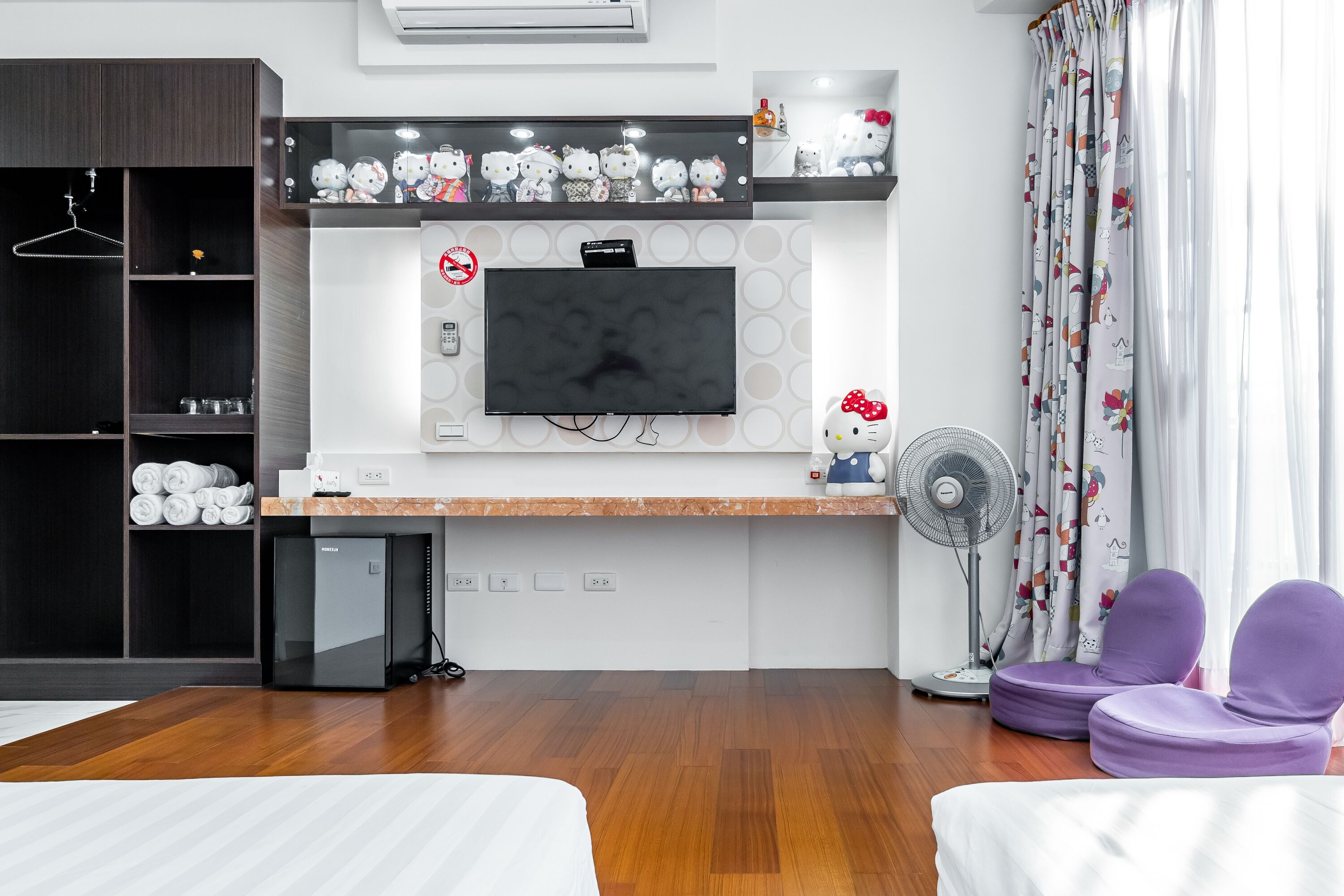 A modern hotel room with a flat-screen TV, a minibar, a small fridge, a fan, and a bed.