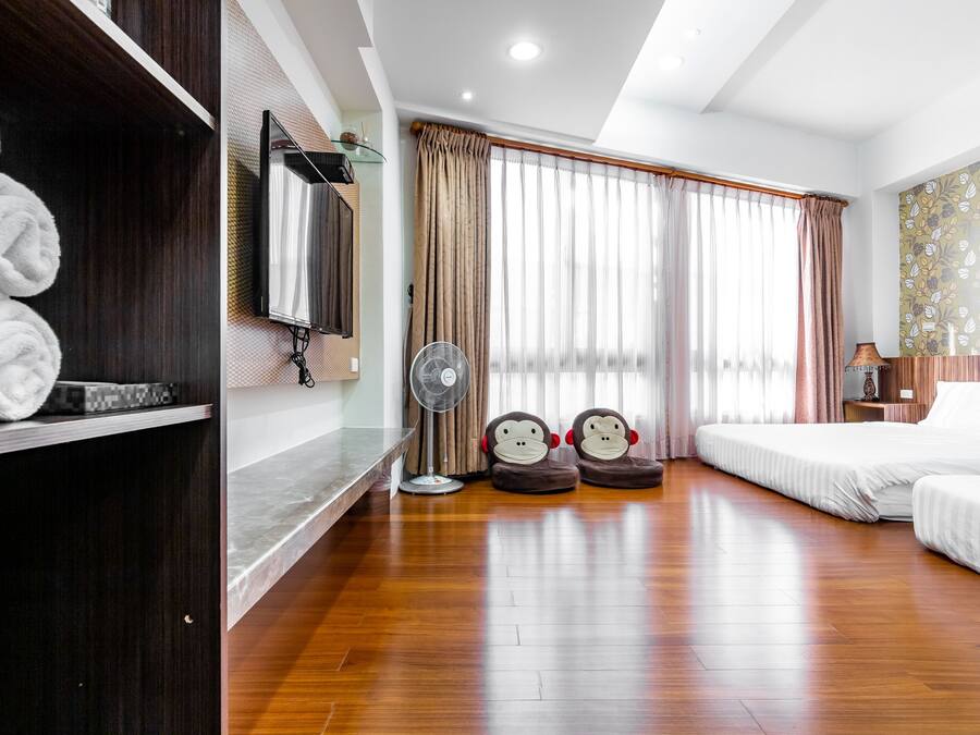 A hotel room with a wooden floor, a bed, a desk with a chair, a mirror, a TV, and a window with curtains.