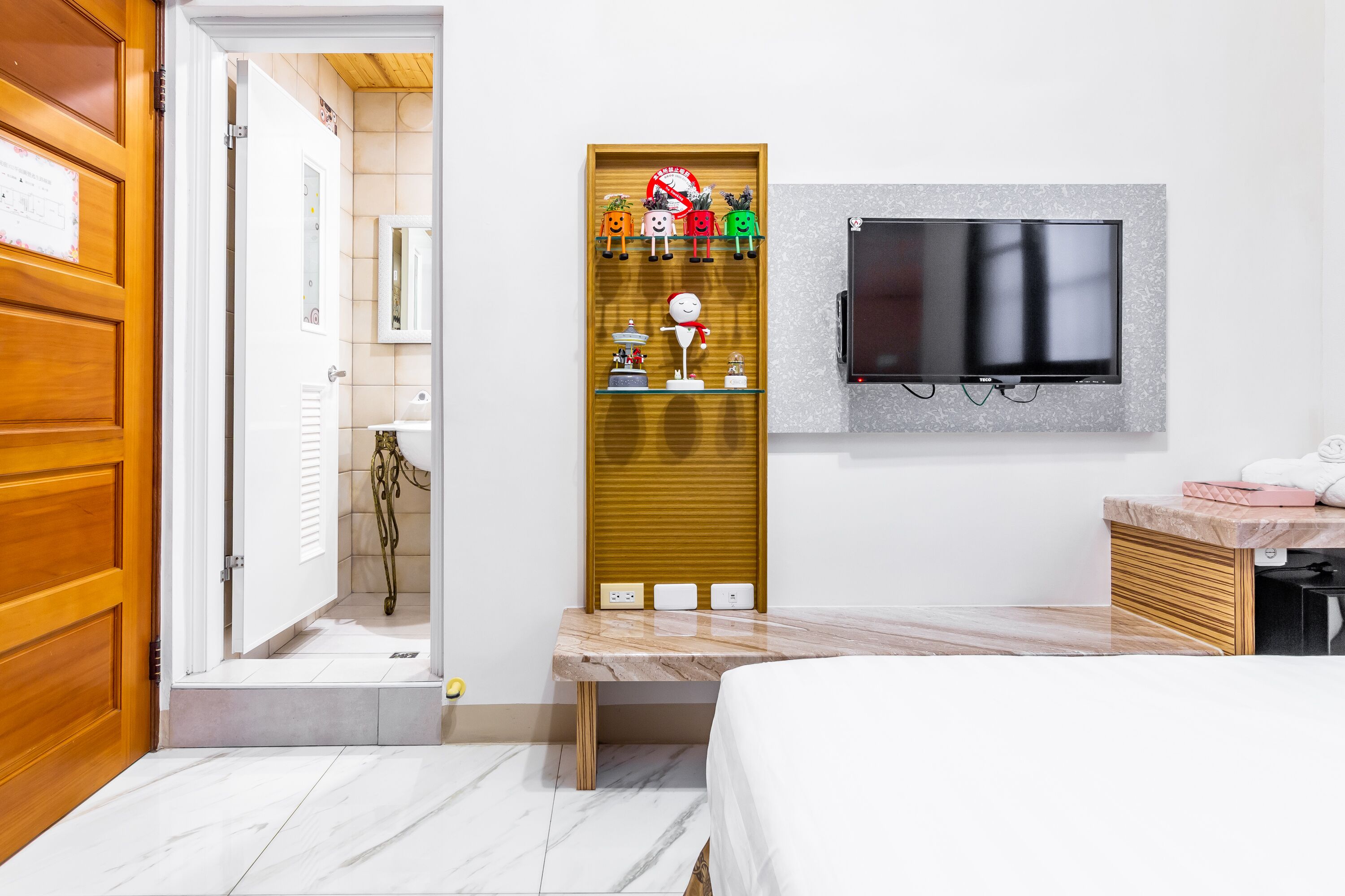 A hotel room with a bed, a TV mounted on the wall, a wooden door, a bathroom with a mirror, and a shelf with decorative items.