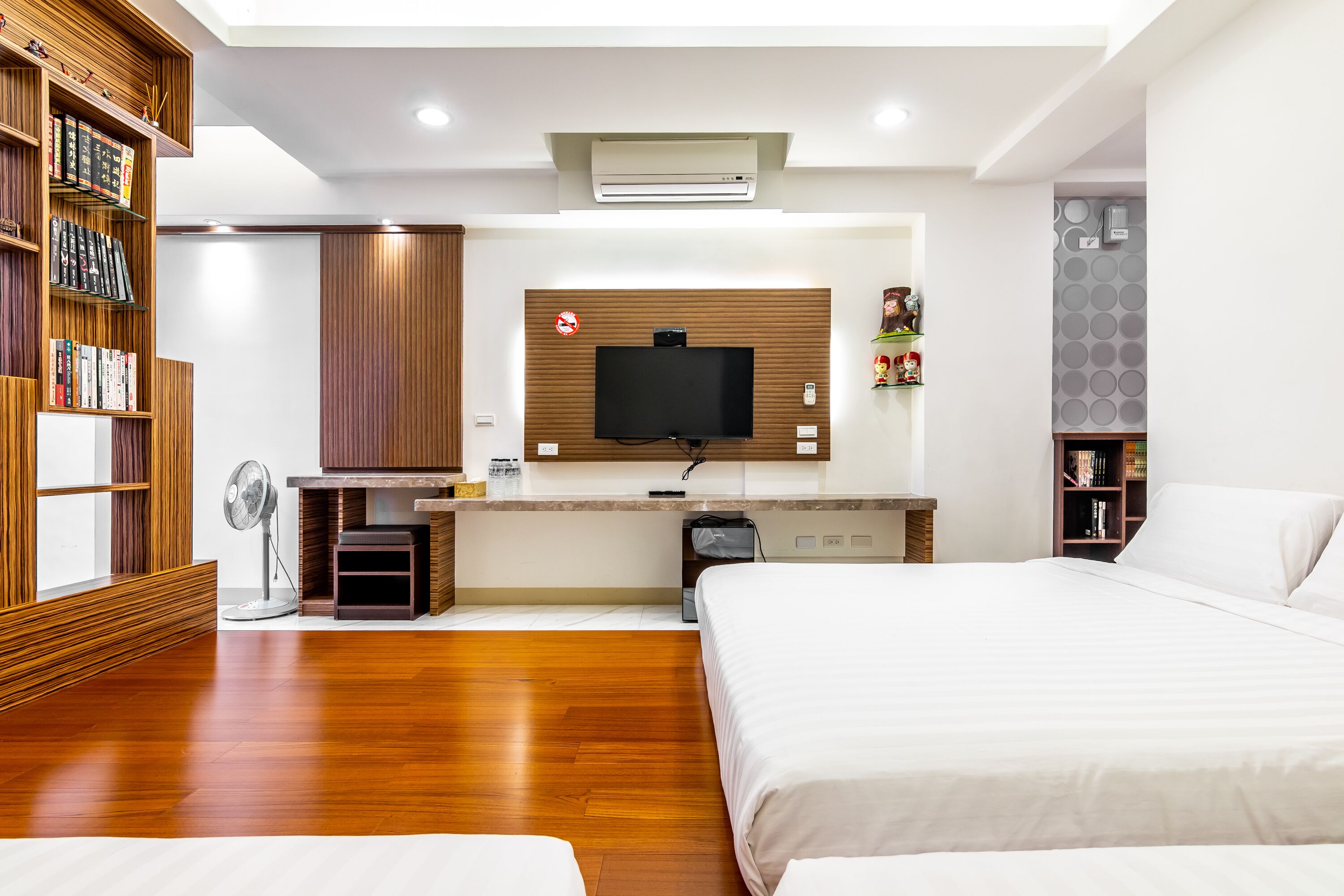 Une chambre d’hôtel moderne avec un plancher en bois, un lit, un téléviseur fixé au mur, qui est recouvert de panneaux en bois, un petit bureau avec une chaise et une bibliothèque.