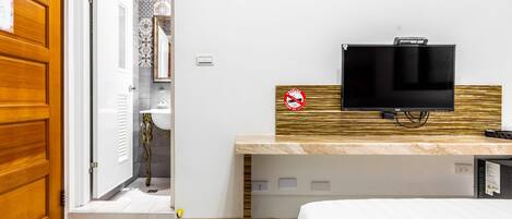 Quarto de hotel com cama, televisão presa a um painel de madeira, banheiro com chuveiro e porta que dá para outro cômodo.