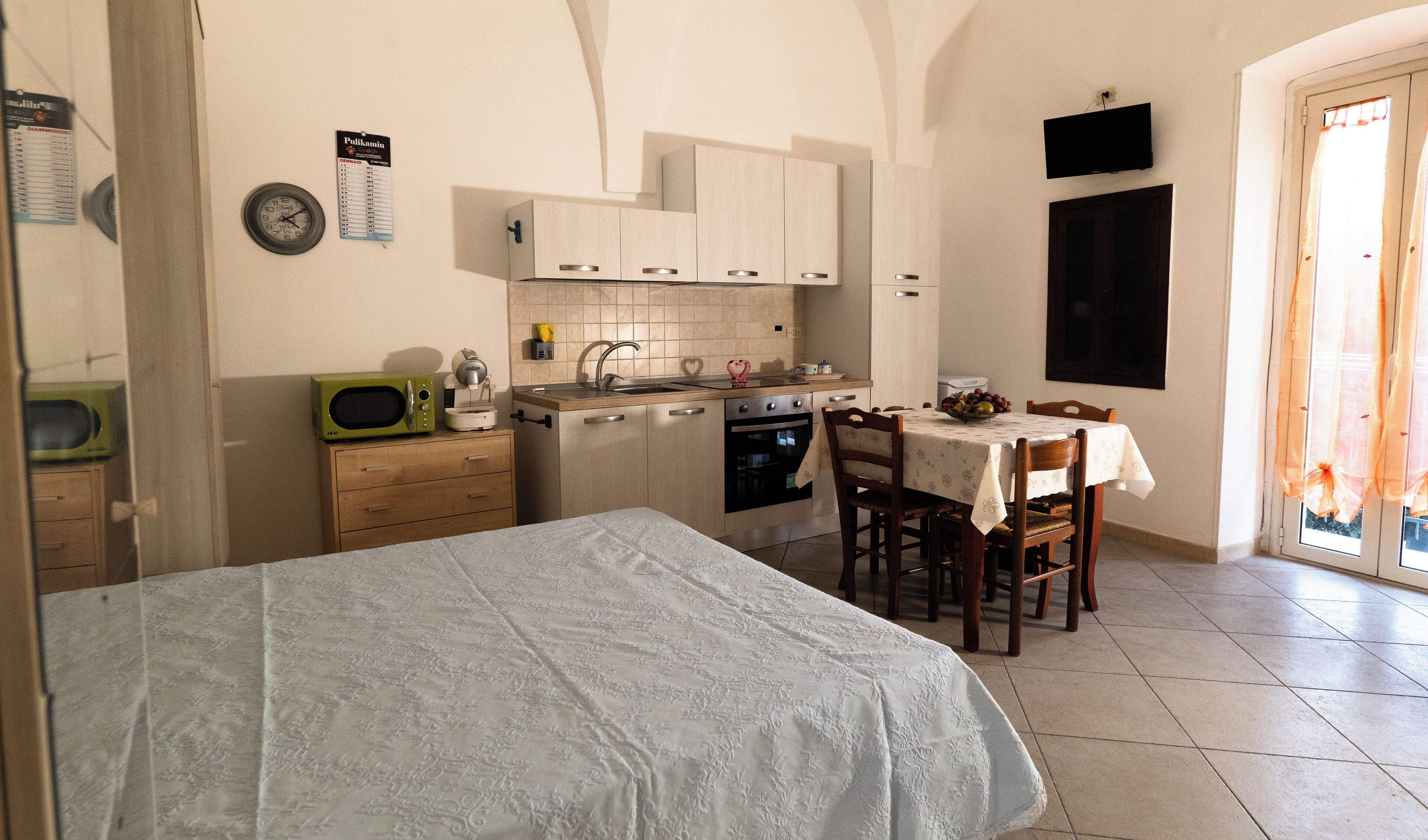 Eine kleine Küche mit Mikrowelle, Backofen und Spüle. Ein Esstisch mit Stühlen und ein Bett mit weißer Bettdecke.
