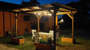 Une terrasse couverte, agrémentée d’une table et de chaises, entourée de jardinières et surplombée d’une pergola en bois illuminée de guirlandes lumineuses.