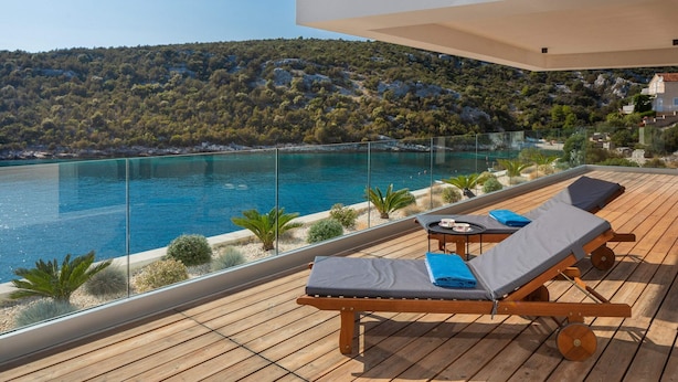 A wooden deck with two lounge chairs and a table, overlooking a clear blue sea and a hilly landscape. - Beachfront Villa Eleganza with Pool (Marina)
