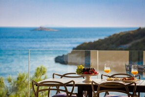 Une table avec un panier de fruits, un croissant et des verres de jus d’orange, donnant sur une vue sur la côte.