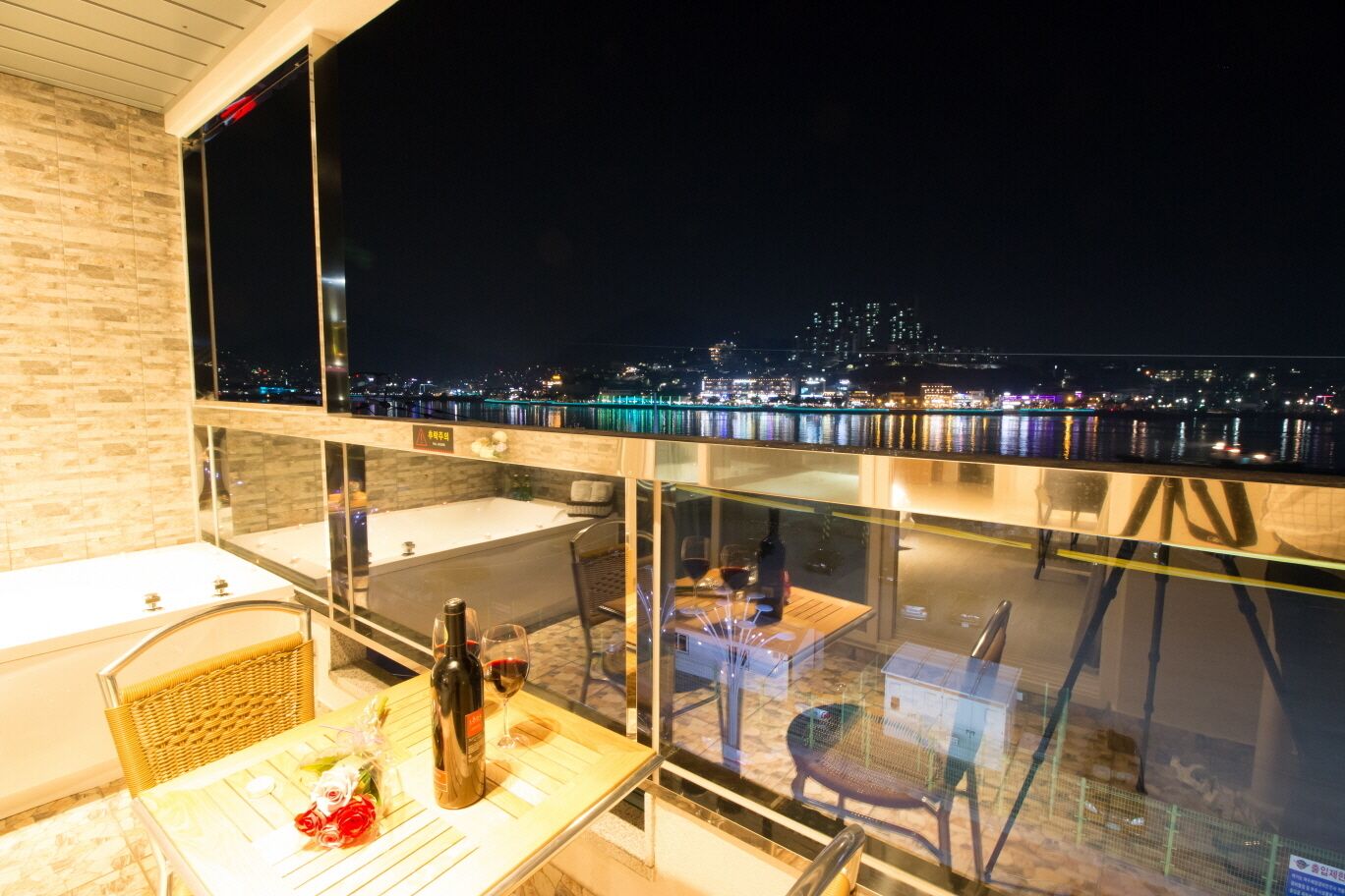 A balcony with a glass barrier, a table with a bottle and glasses, and a view of a cityscape at night.