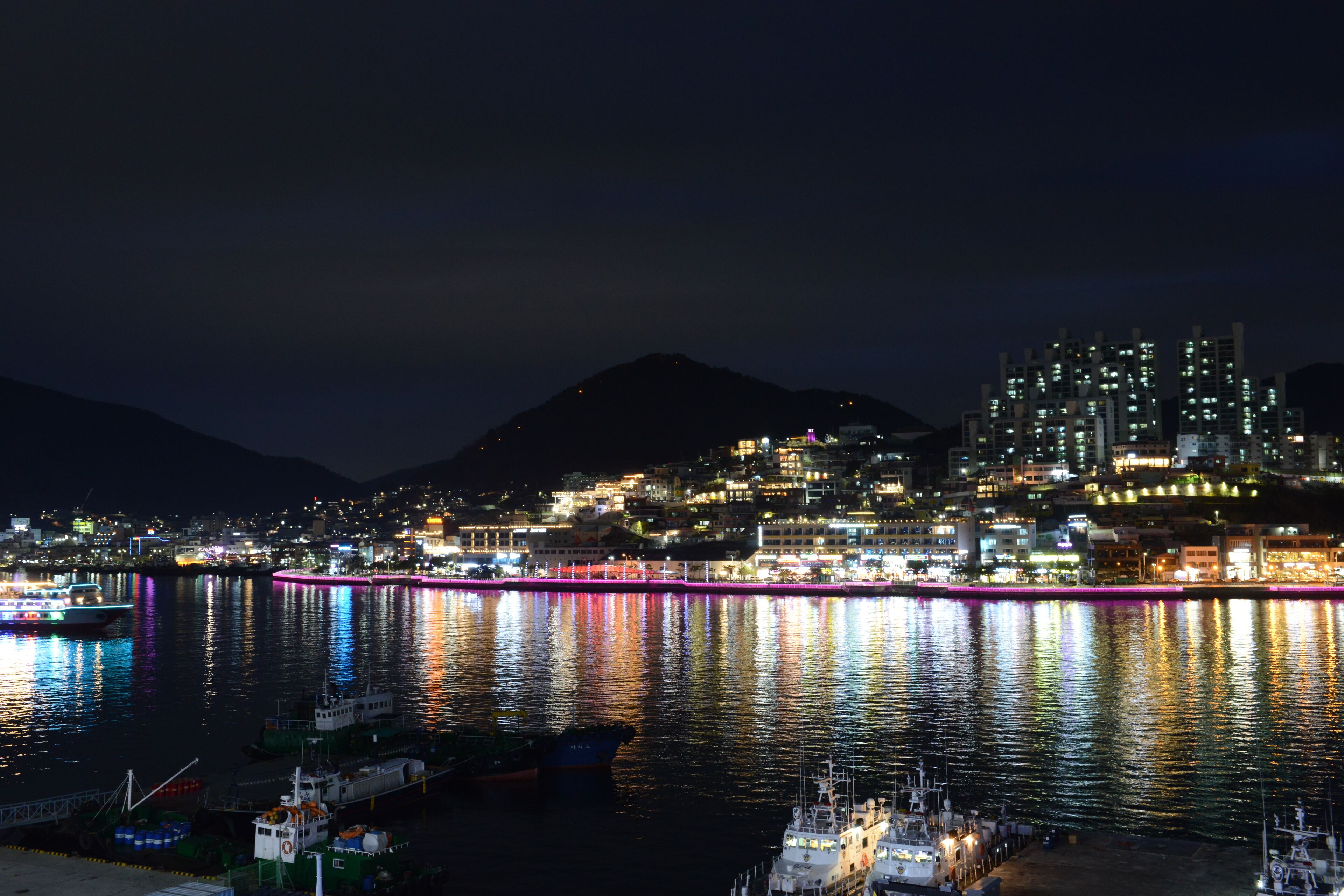 Une vue nocturne d’une ville, avec un front de mer illuminé, des bateaux amarrés et des bâtiments dont les fenêtres sont éclairées.