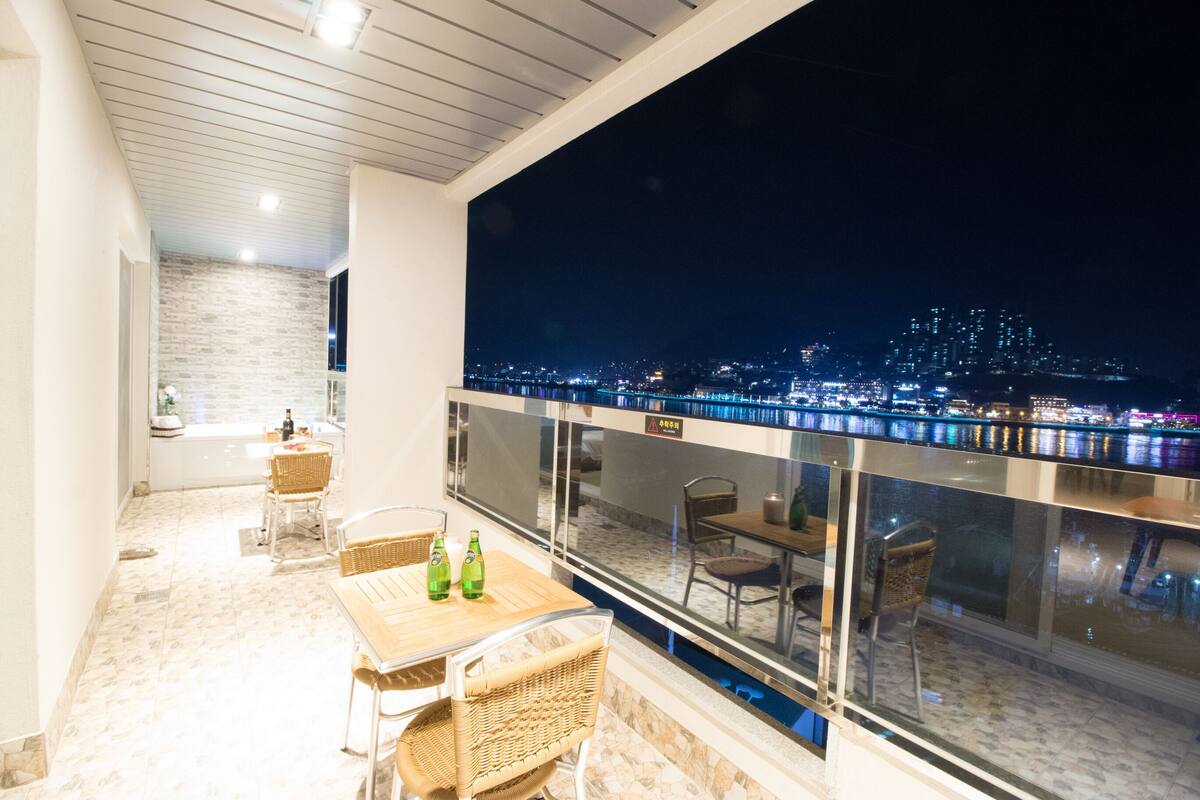 Un balcón con vista a la ciudad de noche, con una mesa, sillas y botellas.