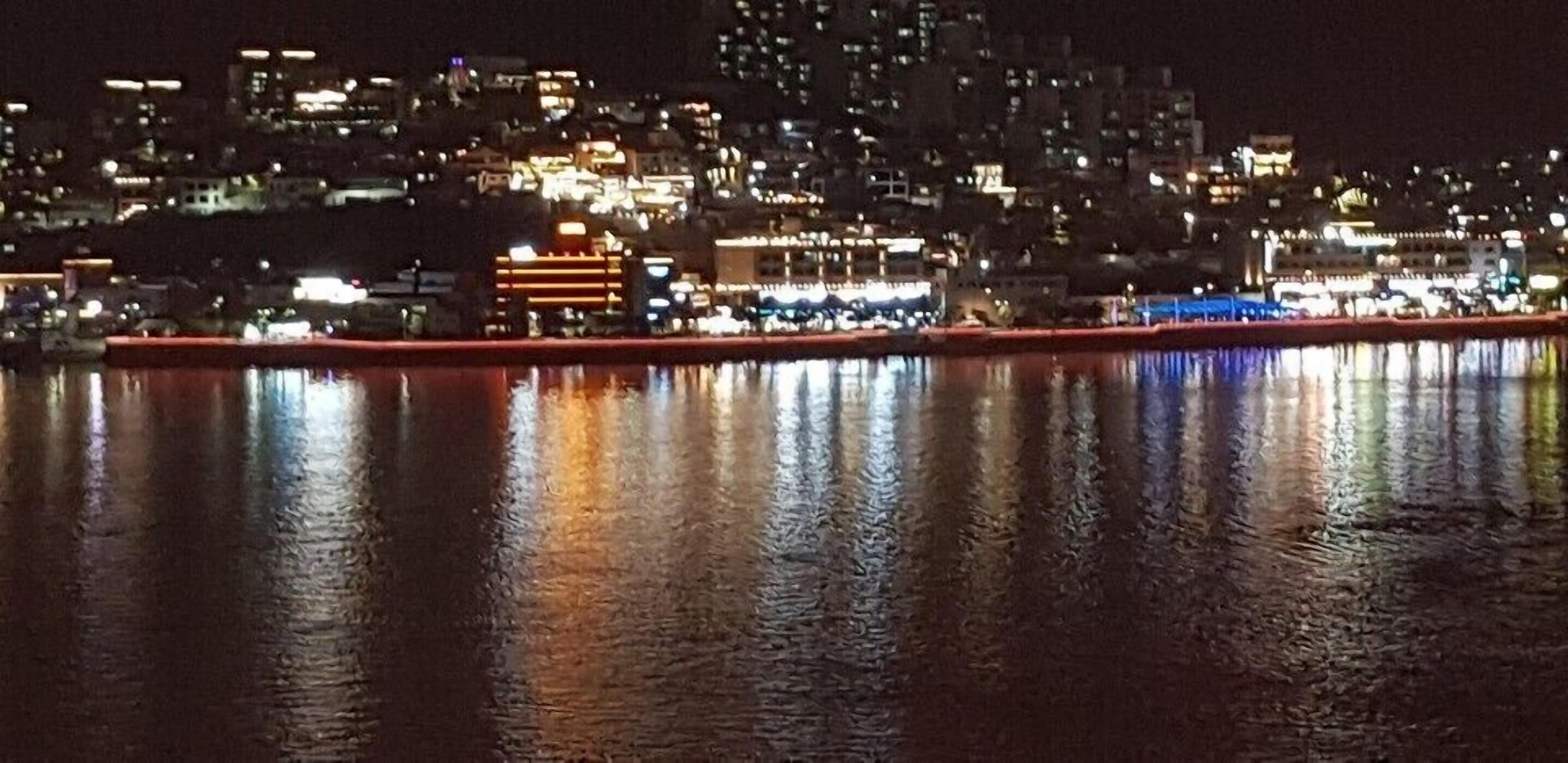 Une vue nocturne d’une ville, avec les bâtiments qui se reflètent dans l’eau calme.