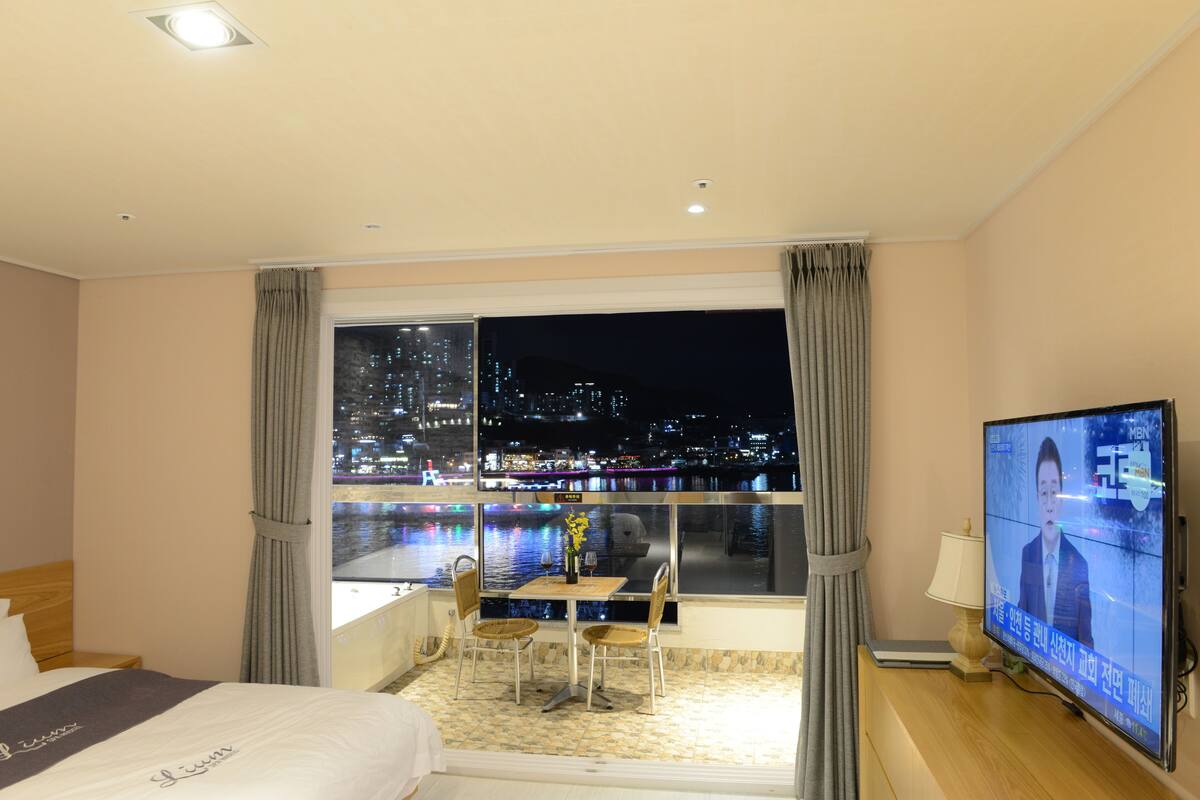 Habitación de hotel con cama, televisión, zona de comedor con sillas y mesa, y vista a un paisaje urbano nocturno.