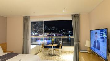 A hotel room with a bed, a TV, a dining area with chairs and a table, and a view of a cityscape at night.