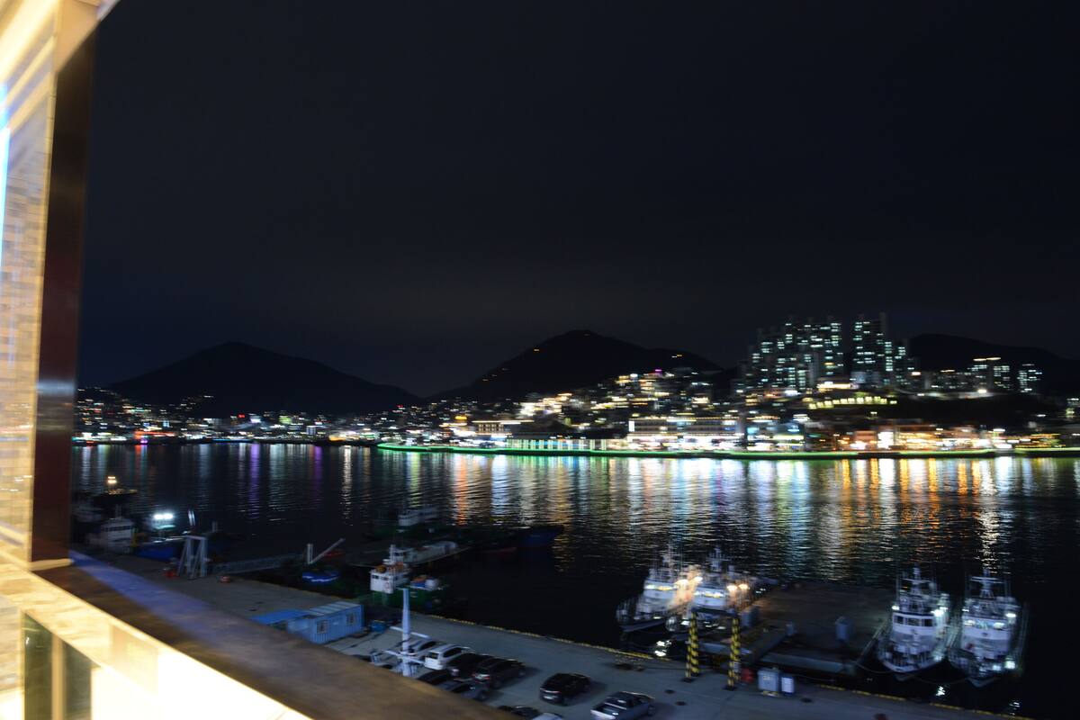 Vista nocturna de un paisaje urbano frente al mar, con edificios iluminados y barcos amarrados en un puerto deportivo.