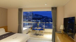 Habitación de hotel con cama, escritorio con lámpara, silla, mesita con un jarrón y vista a un paisaje urbano nocturno.