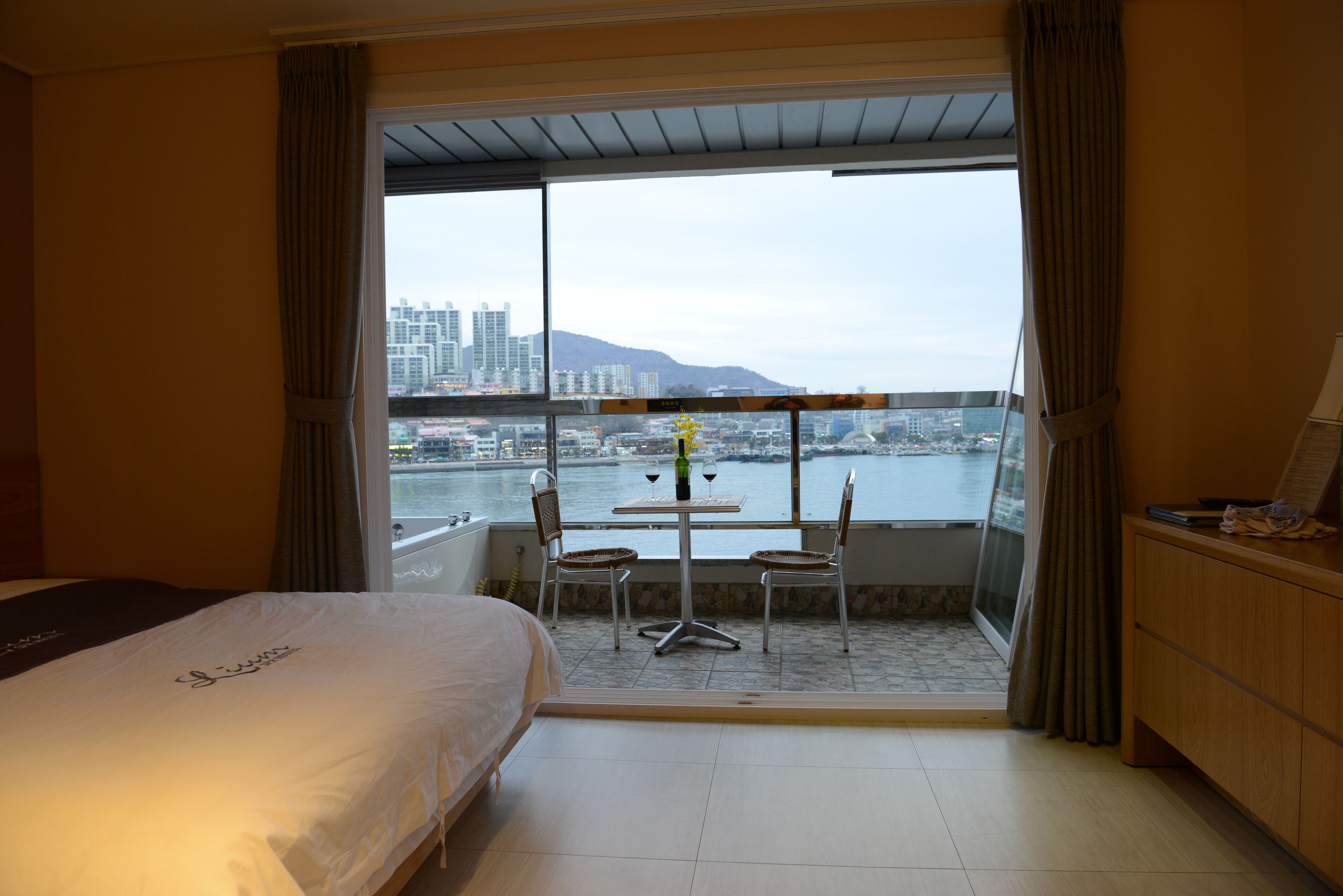 Une chambre d’hôtel avec un balcon, un lit, une table avec des chaises et une vue sur le paysage urbain et l’eau.