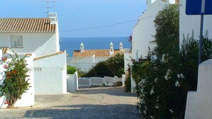 Une rue pavée mène à un bâtiment blanc, d’où l’on a vue sur la mer, et qui est bordée d’une clôture blanche.