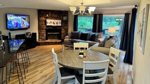 A cozy living room with a stone fireplace, a television, a dining table with chairs, and a large window with curtains.