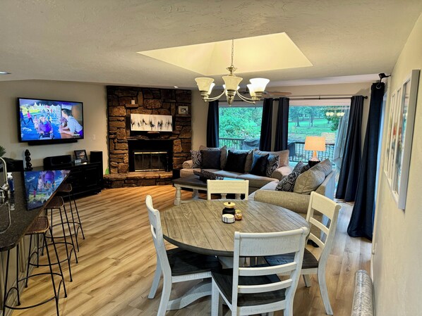A cozy living room with a stone fireplace, a television, a dining table with chairs, and a large window with curtains.