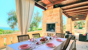 An outdoor dining area with a stone fireplace, a table set for four with pink plates, and a view of the sea. - Cappero Pool House by Galatea Holiday Homes (Gagliano del Capo)