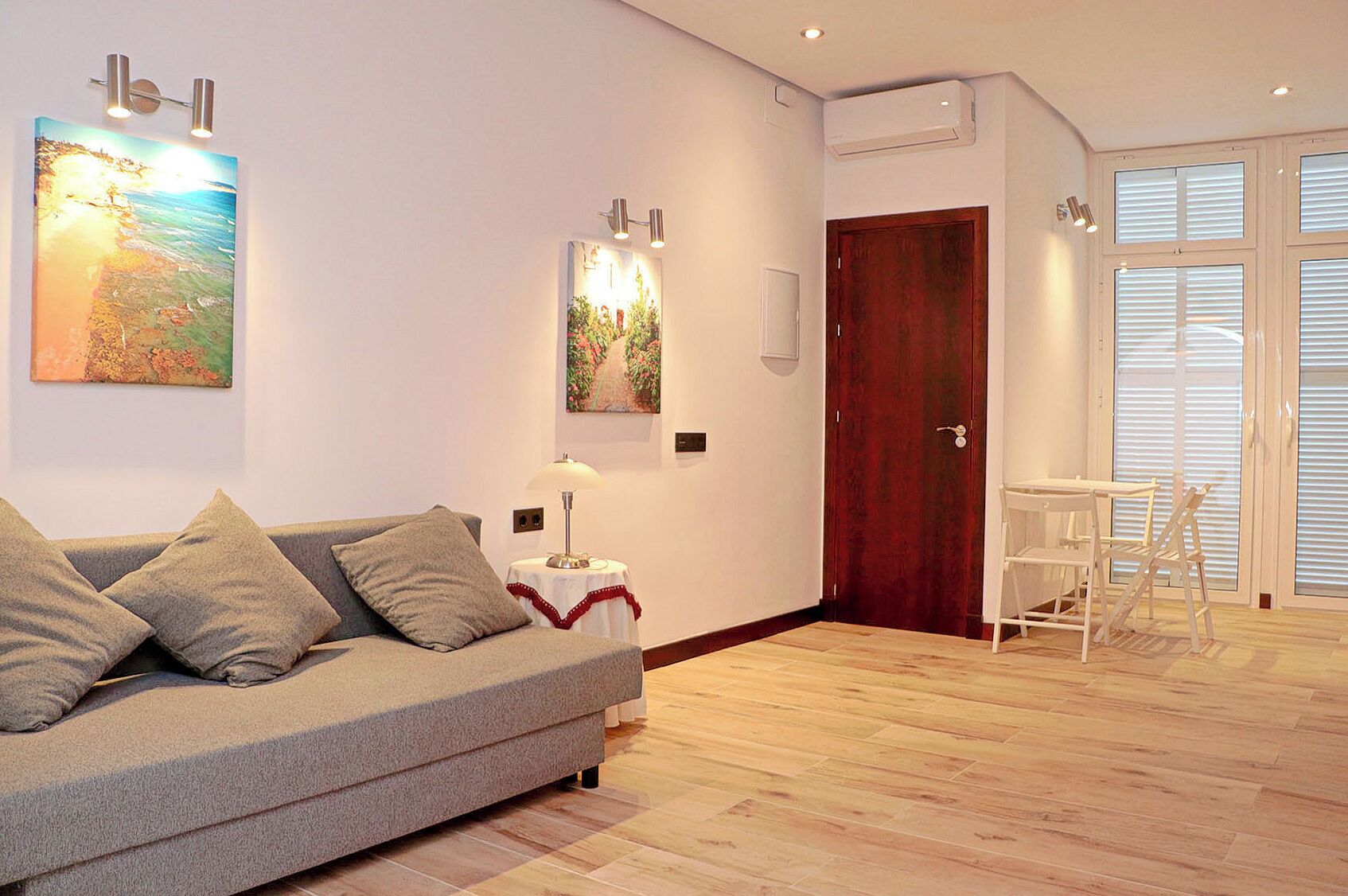 A modern living room with a grey sofa, a small table with a lamp, and a wooden floor. There are two paintings on the wall and a wooden door in the background.