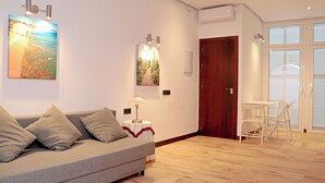 A modern living room with a grey sofa, a small table with a lamp, and a wooden floor. There are two paintings on the wall and a wooden door in the background.