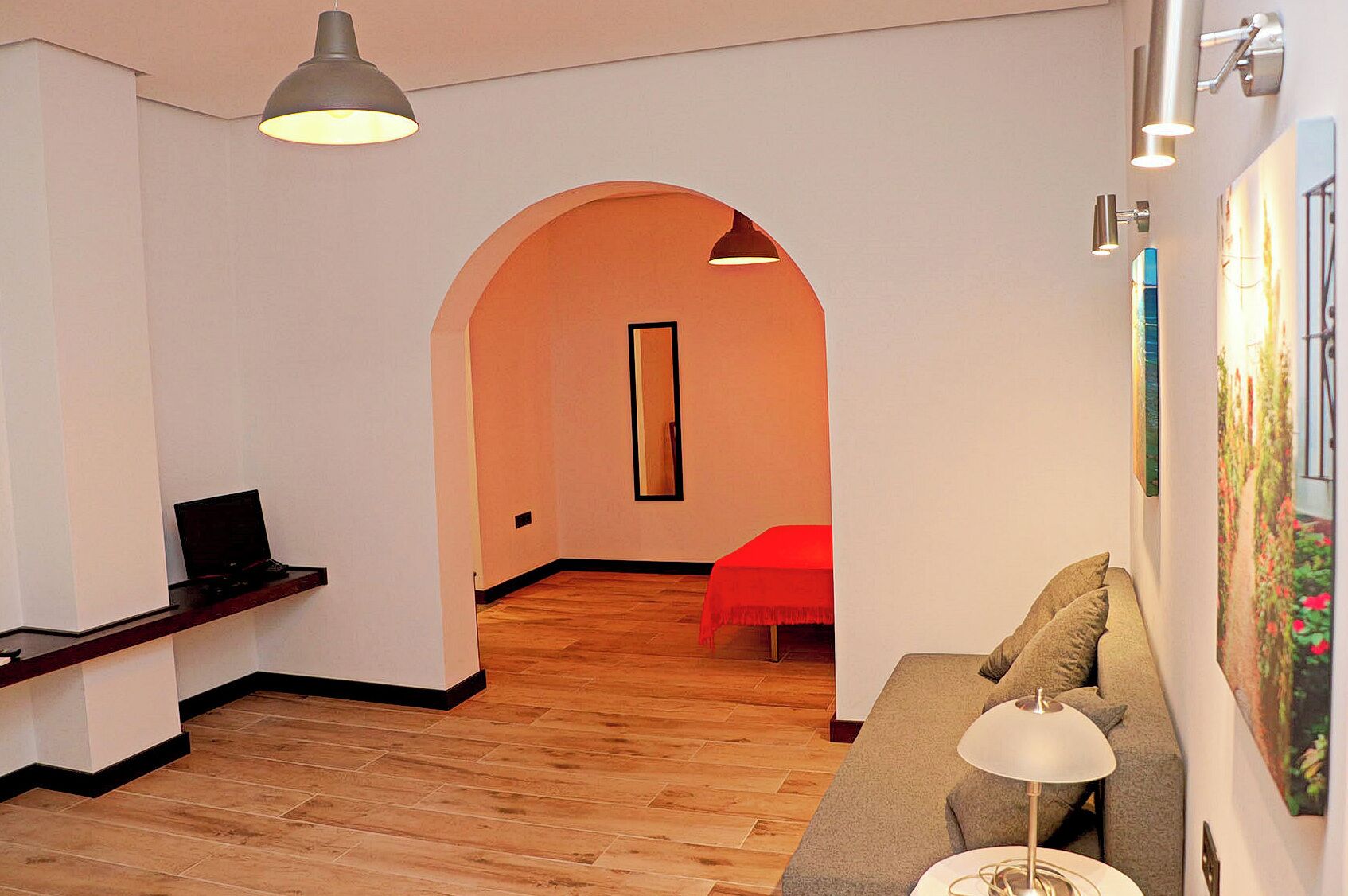 A modern living room with a wooden floor, a sofa, a red chair, a desk with a laptop, and a large painting on the wall.