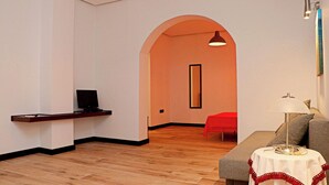 A modern hotel room with a wooden floor, a grey sofa, a small round table with a white tablecloth, a red bed, and a TV on a wooden shelf.