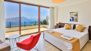 A hotel room with a large bed, a red chair, a small table, and a view of the sea and mountains.